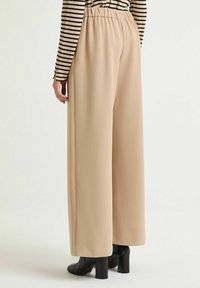 Pantaloni beige a gamba larga con una struttura liscia e una cintura elastica, abbinati a stivaletti neri con texture. Camicia a maniche lunghe a righe visibile.