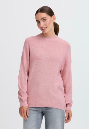 Maglione rosa a maglia con maniche lunghe e collo alto, caratterizzato da una trama a costine e un taglio rilassato. Abbinato a jeans grigi chiari.