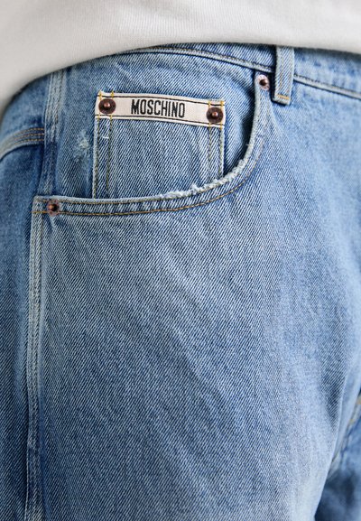 Ljusblå denimjeans med en sliten textur, med en Moschino-logotyp på bakfickan och kopparnitar för förstärkning.