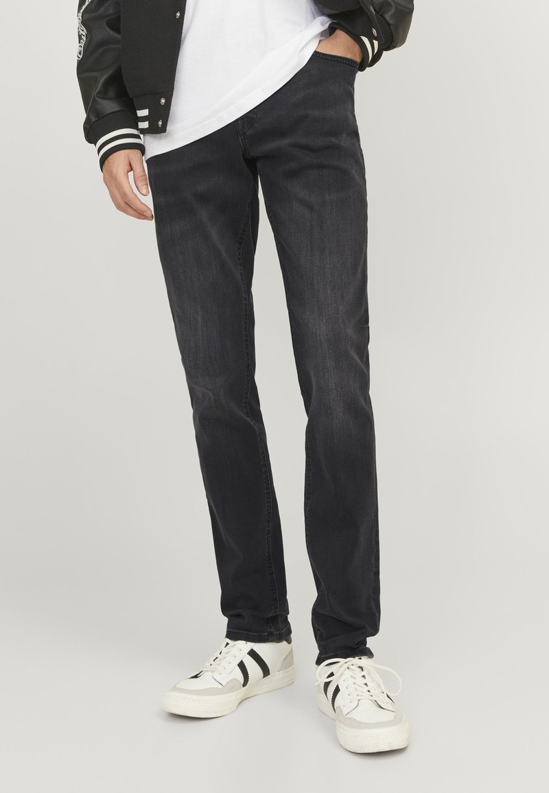 Uomo che indossa jeans neri, sneakers bianche con strisce nere, camicia bianca e giacca nera con bordo bianco, in piedi con una mano in tasca.