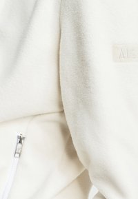 Veste en polaire blanche avec une texture lisse, une poche zippée et un logo discret de couleur beige clair.