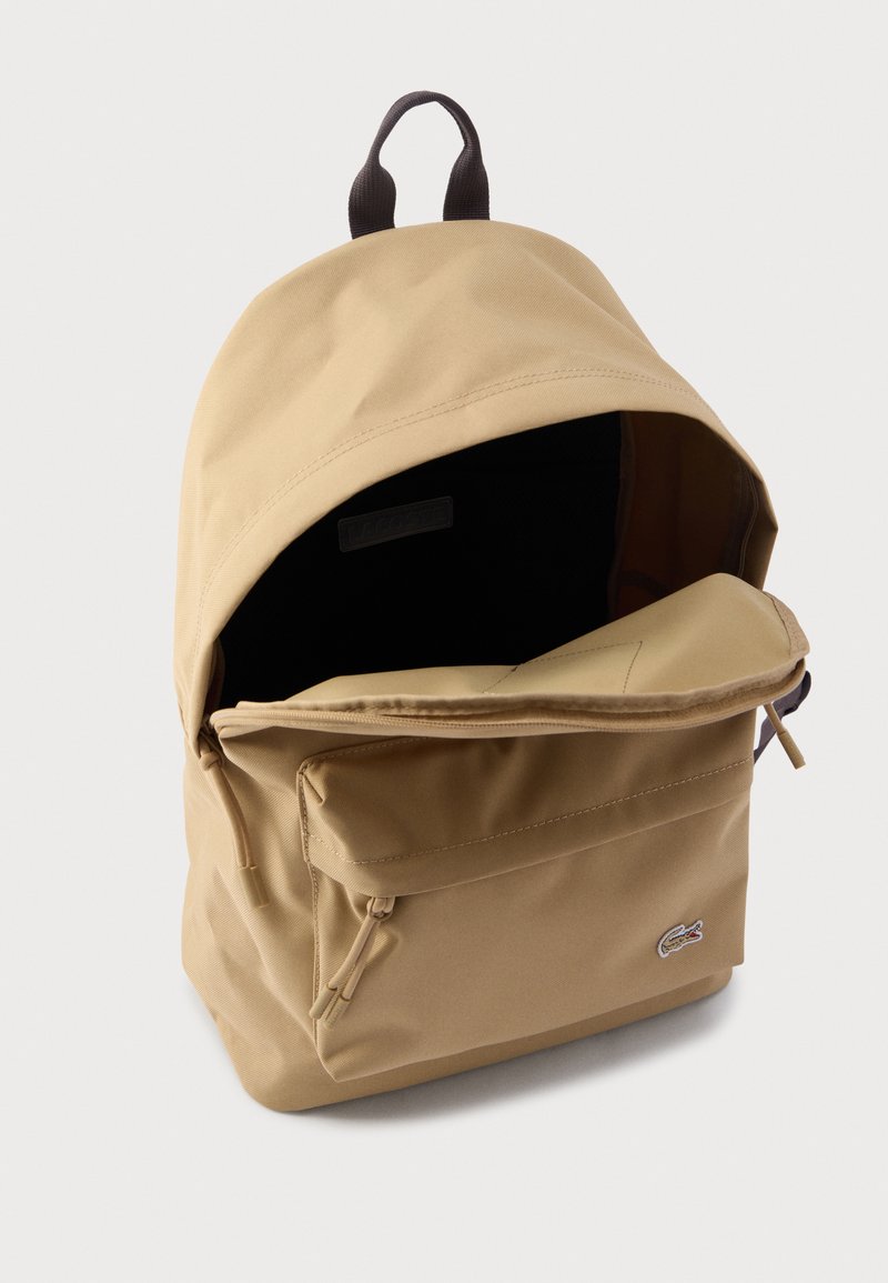 Mochila beige con tela texturizada, que cuenta con un bolsillo frontal con cremallera y una asa superior. El interior es negro, con un logo de Lacoste en el lateral.
