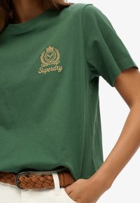 T-shirt vert à col rond en coton doux, avec un logo brodé en or et un motif de feuille sur la partie gauche de la poitrine.