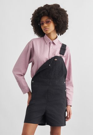 Vrouw met afro-kapsel die een roze textuur lange mouwen shirt draagt, zwarte denim korte overalls en getinte rechthoekige zonnebril.