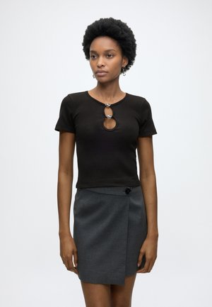 Νεαρή γυναίκα με κοντά σγουρά μαλλιά που φοράει μαύρο τοπ με κοντά μανίκια και λεπτομέρεια κλειδαρότρυπας και γκρι φούστα τύπου ζώνη, στέκεται μπροστά σε απλό φόντο.