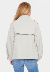 Jachetă supradimensionată gri deschis cu un design cu guler pliat, având un guler clasic și accente cu butoane pe mâneci. Purtată cu blugi albaștri.