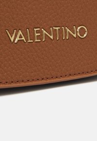 Brunt lädermaterial med en texturerad yta, prytt med en gyllene "VALENTINO"-logotyp. Synlig söm längs kanten lägger till detaljer.