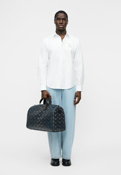 Άνδρας που φοράει λευκό πουκάμισο με κουμπιά και ανοιχτό μπλε τζιν, κρατώντας μαύρη τσάντα ταξιδιού με σχέδια από τις λαβές της, στέκεται μπροστά σε λευκό φόντο.