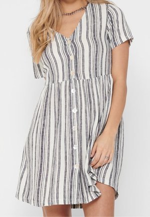 Frau trägt ein kurzärmeliges, schwarz-weiß gestreiftes, knöpfbares Kleid mit V-Ausschnitt und Kettenhalskette, Hand ruht am Saum des Rocks.