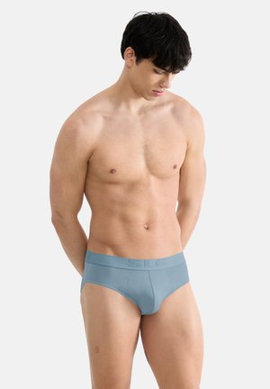 Nuori mies, jolla on tummat hiukset ja vaaleansiniset alushousut, seisoo käsivarret selän takana ja katsoo alas yksiväristä taustaa vasten.