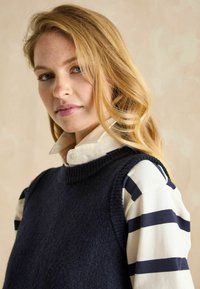 Marineblau gestrickte Weste über einem langärmligen Oberteil in Weiß und Marineblau gestreift. Der Ausschnitt ist rund und der Stoff hat eine weiche Textur.