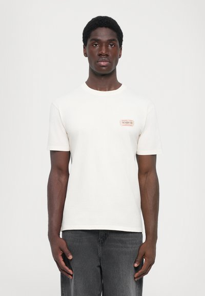 T-shirt crème clair à manches courtes, col rond, avec un petit détail graphique rectangulaire sur la poitrine gauche. Le tissu semble doux.