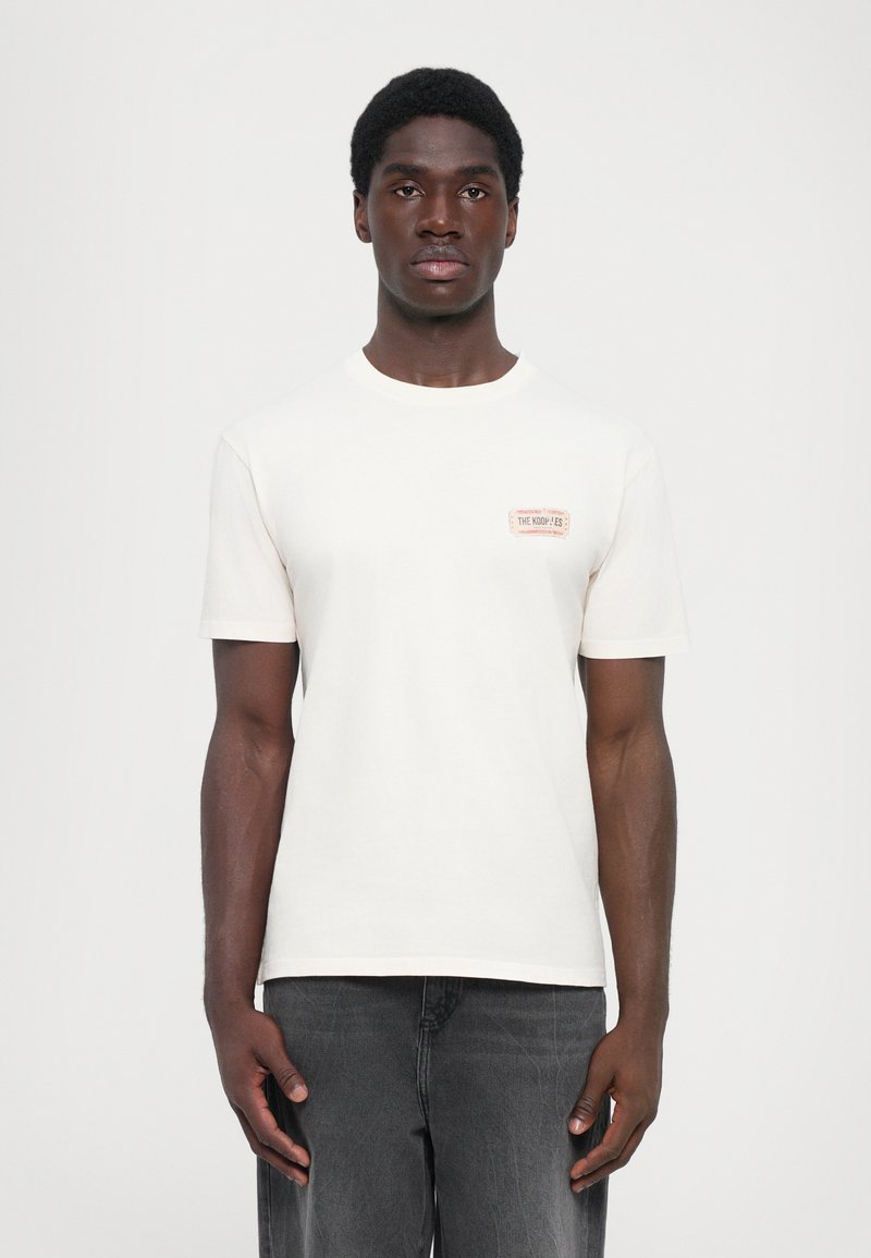 Light cream t-shirt with short sleeves, round neck, and a small, rectangular graphic detail on the left chest. Fabric appears soft.