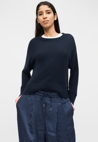 Γυναίκα που φοράει ένα χαλαρό πουλόβερ σε ναυτικό μπλε χρώμα πάνω από λευκό τοπ και ναυτικά μπλε παντελόνια με πουά και κορδόνι, στέκεται με τα χέρια στις τσέπες.