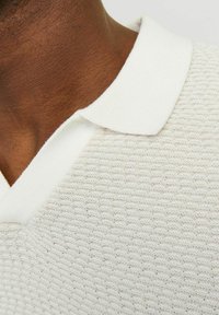 Bílá texturovaná polo košile s ribbovaným límcem, zobrazující vzor včelí plástve a měkkou látku. Detailní pohled zaměřený na detaily výstřihu.