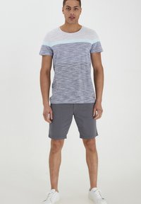T-shirt med korte ærmer og rund halsudskæring har et blåt og gråt stribet mønster, båret med grå shorts og hvide sneakers på en ensfarvet baggrund.