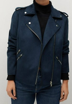 Kvinde iført en mørkeblå ruskind bikerjakke med sølv lynlåse og knapper over en sort rullekravesweater og blå jeans.
