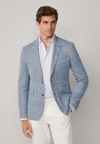 Hellblaues Tweed-Blazer mit zwei Fronttaschen, Reverskragen und dunklen Knöpfen. Getragen über einem weißen Hemd mit Kragen und beigen Hosen.