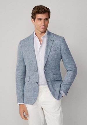 Blazer en tweed bleu clair avec deux poches avant, revers crantés et boutons foncés. Porté sur une chemise blanche à col et un pantalon beige.