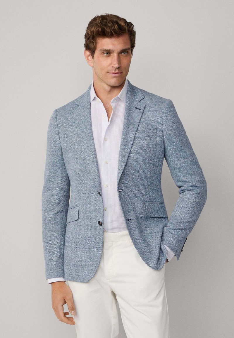 Hellblaues Tweed-Blazer mit zwei Fronttaschen, Reverskragen und dunklen Knöpfen. Getragen über einem weißen Hemd mit Kragen und beigen Hosen.