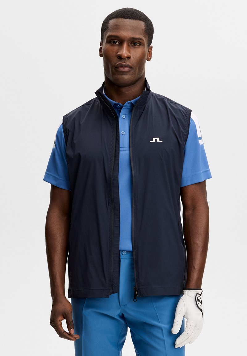 Mand iført en mørkeblå ærmeløs vest over blåt golftøj med en hvid golfhandske på højre hånd mod en ensfarvet baggrund.