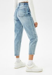 Henkilö, jolla on päällään vaaleansiniset korkeavyötäröiset lyhyet farkut, valkoinen ribbipintainen pitkähihaisuus, valkoiset sukat ja valkoiset korkeat lenkkarit, joissa on kultaketjuviimeistely.