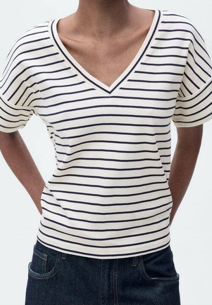 Personne portant un t-shirt blanc à col en V avec des rayures horizontales noires et un jean en denim foncé, les mains dans le dos.