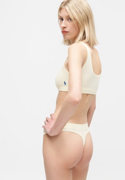 Femme blonde portant un soutien-gorge crème et une culotte string assortie, posant avec la main sur la hanche, regardant par-dessus son épaule sur un fond uni.