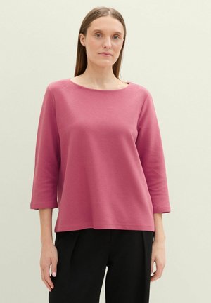 Haut à manches longues rose en tissu texturé, avec un décolleté large et une coupe ample. Associé à un pantalon noir.