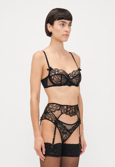 Ensemble de lingerie en dentelle noire avec des motifs floraux, texture transparente, bretelles réglables et nœuds décoratifs, associé à des bas autoportants.