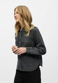 Chemise en denim gris foncé avec des boutons, manches longues et un ourlet courbé. Présente des coutures contrastées et une coupe décontractée.