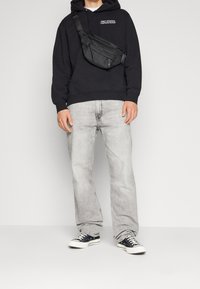 Černá mikina s kapucí a bílým nápisem, světle šedé džínové kalhoty, černá kabelka přes rameno a černé tenisky s bílými detaily, stojící na neutrálním pozadí.