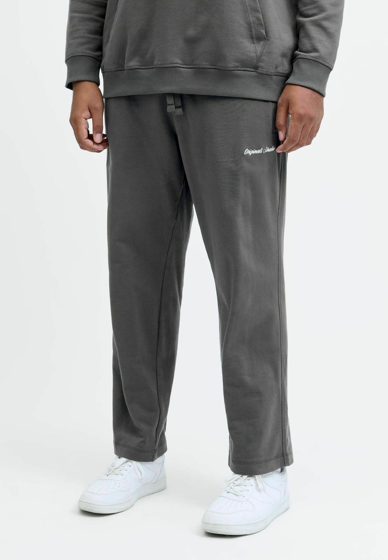 Osoba ubrana w ciemnoszare spodnie dresowe z napisem "Originals Studio", pasującą bluzę oraz białe trampki, stojąca na białym tle.