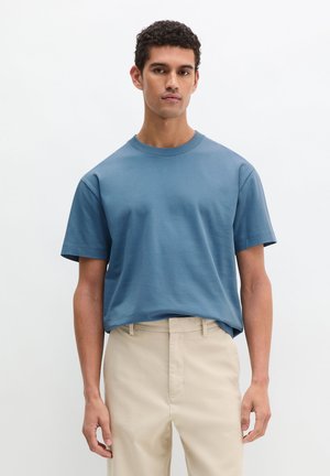 Mannelijk model draagt een effen blauw T-shirt met korte mouwen, ingestopt in beige broek, staand tegen een eenvoudige witte achtergrond.