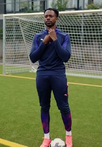 Sportininkas tamsiai mėlyname Puma treninge su purpuriniais akcentais, stovintis ant žalios žolės, laikantis apykaklę, dėvintis rožinius sportinius batelius ir turintis futbolo kamuolį po kojomis.