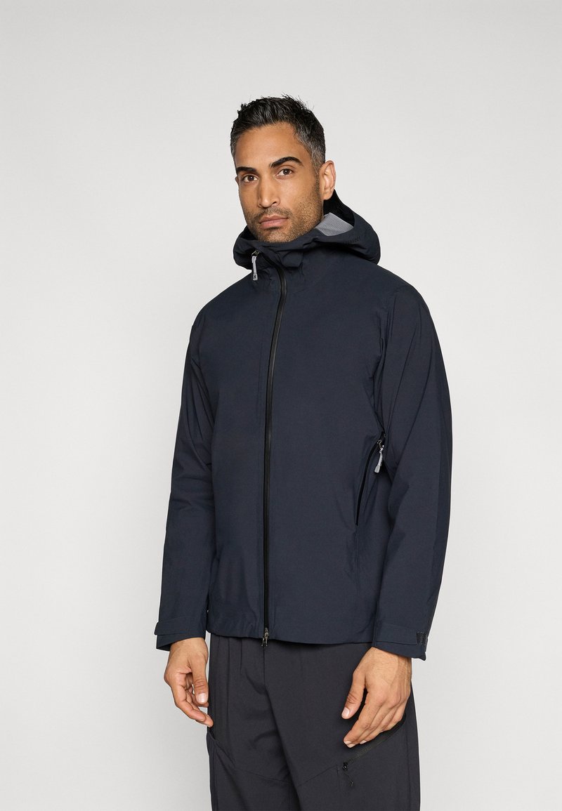 Marineblå vandtæt jakke med lynlås foran, justerbar hætte, sidelommer med lynlås og elastiske manchetter; glat tekstur og figursyet design.