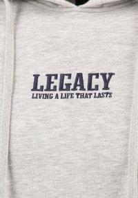 Lichtgrijze hoodie gemaakt van zachte, gestructureerde stof. Voorzien van donkerblauwe geborduurde tekst: "LEGACY LIVING A LIFE THAT LASTS." Het hoodie-ontwerp bevat een voorzak.