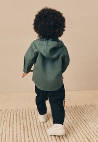 Hanorac verde cu glugă, design casual, combinat cu pantaloni cargo negri cu buzunare laterale și o etichetă cu logo. Pantofii sport albi completează ținuta.