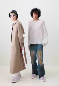 Casaco trench bege, suéter leve, denim desgastado e ténis. Acessórios notáveis incluem óculos de sol e um chapéu preto.