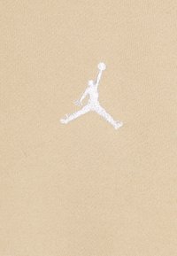 Tessuto beige con una trama intrecciata, caratterizzato da un logo Jumpman ricamato in bianco al centro. Il design è semplice e pulito.