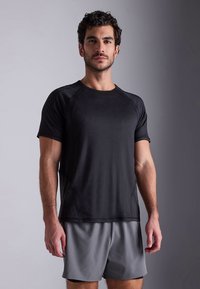 Svart treningsskjorte med korte raglanermer, kombinert med grå shorts. Jevn tekstur, fukttransporterende stoff og rund hals.