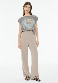 Camiseta gris de manga corta con un gráfico de calavera y rosa, combinada con pantalones de pierna ancha beige. Sandalias negras acentúan el atuendo.