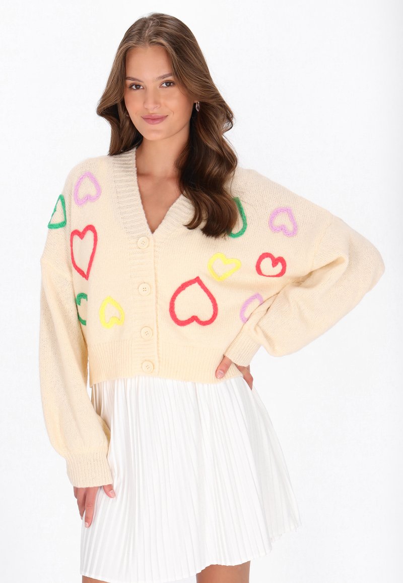 Cardigan crem cu decolteu în V, având modele colorate cu inimi în roșu, galben, verde și violet. Cinci butoane și mâneci tip balon.
