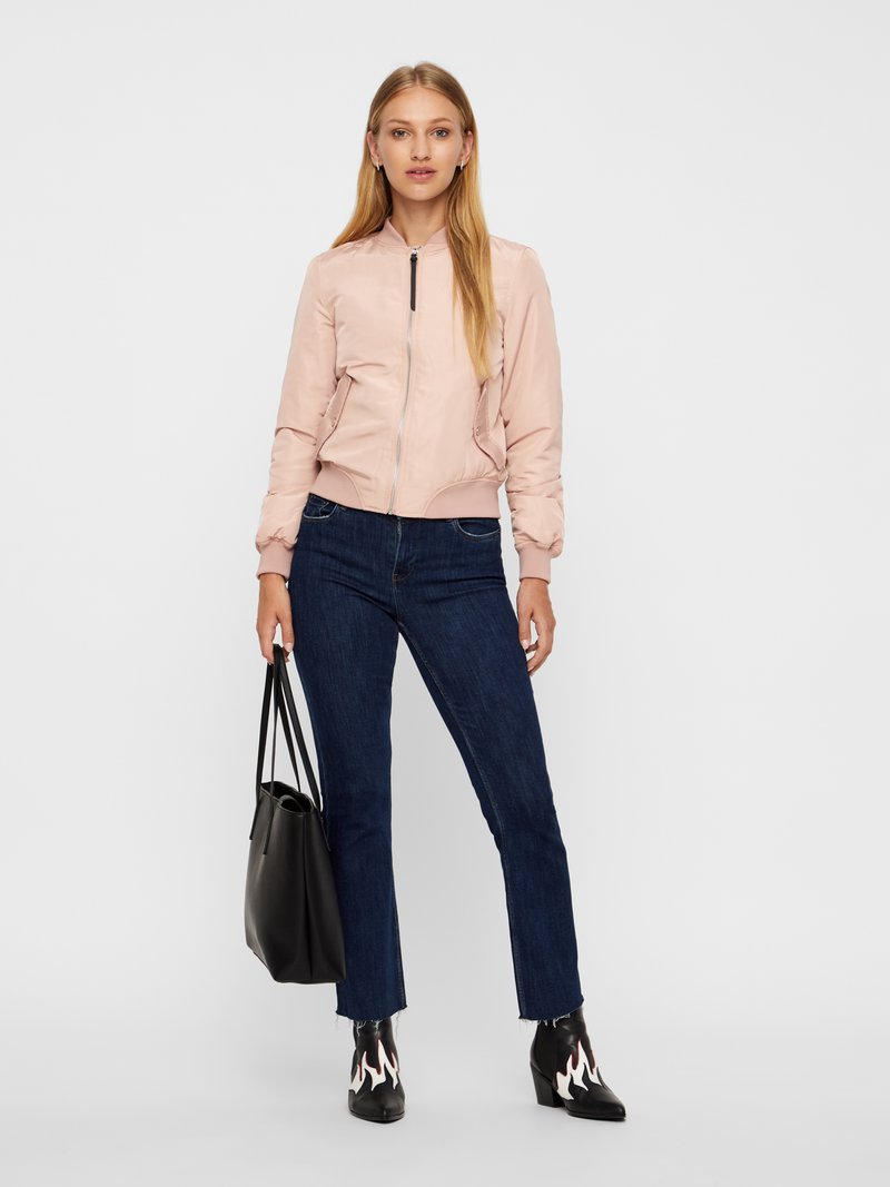 Nainen, jolla on vaaleanpunainen bomber-takki, tummansiniset farkut, mustat liehikkomalliset saappaat ja musta ostoskassi kädessään, seisoo valkoista taustaa vasten.