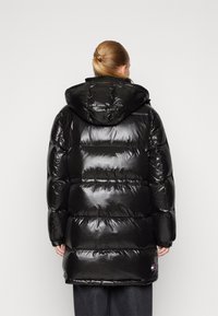 Chaqueta acolchada negra con un exterior brillante, capucha y secciones acolchadas horizontales. Cuenta con puños elásticos y un logo en el bajo.