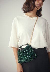 Sac à main vert en sequins avec une forme structurée, doté d'une poignée noire et d'une bandoulière chaîne dorée. Porté avec une chemise blanche oversize.