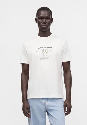 Άνδρας που φοράει λευκό μπλουζάκι Armani Exchange με λογότυπο και ανοιχτό μπλε τζιν, στέκεται μπροστά σε απλό λευκό φόντο.