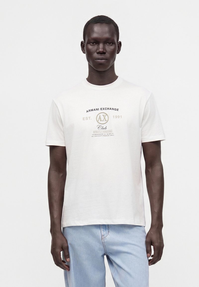 Άνδρας που φοράει λευκό μπλουζάκι Armani Exchange με λογότυπο και ανοιχτό μπλε τζιν, στέκεται μπροστά σε απλό λευκό φόντο.