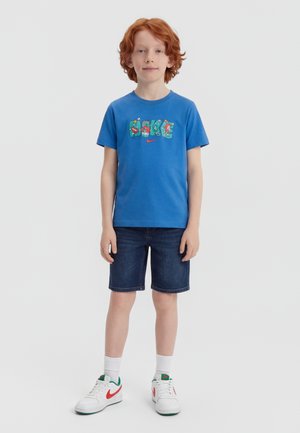 Chlapec s červenými vlasmi stojí v modrom tričku Nike s grafickým motívom, tmavých rifľových kraťasoch, bielych ponožkách a bielych teniskách s červeným logom.