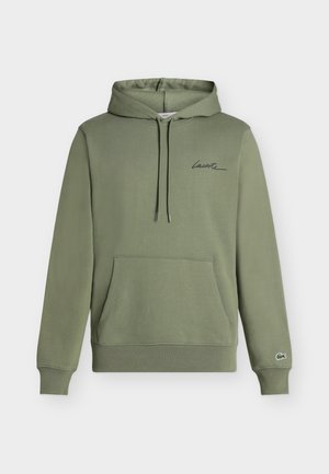 Jeune homme portant un sweat à capuche vert olive, un short noir et des baskets noires, tenant une veste noire pliée sous le bras.